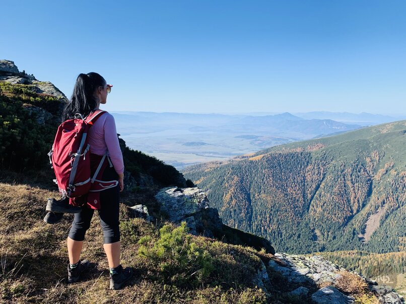 Žena s červeným batohom stojí na vrchole hory a pozerá sa na panorámu dolín a hôr v diaľke.